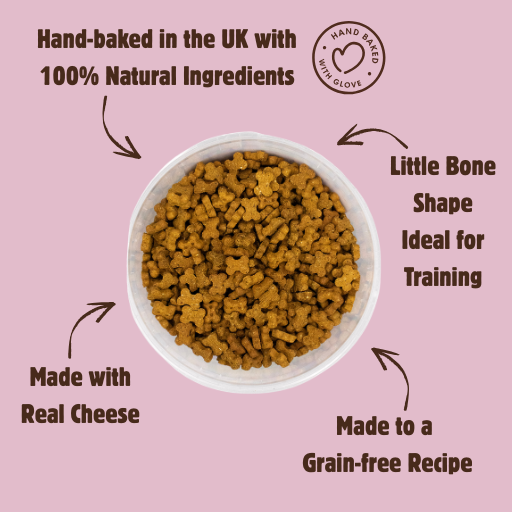 A bowl of small bone-shaped brown dog treats on a dusty pink background with promotional text around it. The text reads: 'Hand-baked in the UK with 100% Natural Ingredients,' 'Little Bone Shape Ideal for Training,' 'Made with Real Cheese,' and 'Made to a Grain-free Recipe.