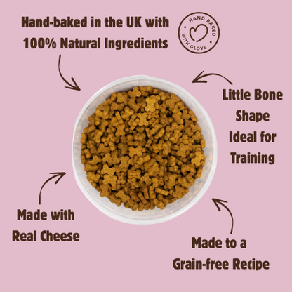 A bowl of small bone-shaped brown dog treats on a dusty pink background with promotional text around it. The text reads: 'Hand-baked in the UK with 100% Natural Ingredients,' 'Little Bone Shape Ideal for Training,' 'Made with Real Cheese,' and 'Made to a Grain-free Recipe.