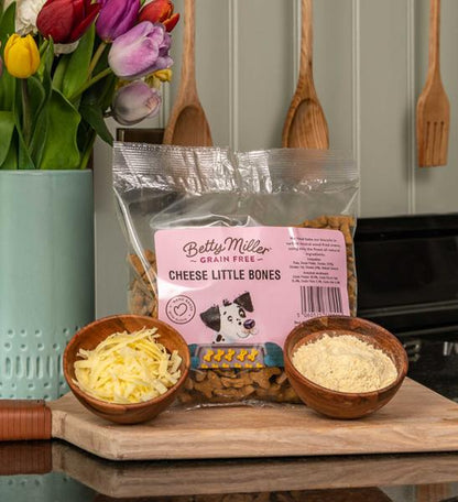 Packaging of 'Betty Miller Grain-Free Cheese Little Bones' with a clear view of the product name, brand, and a picture of the cheese little bones. In the foreground, there are two wooden bowls filled with what appears to be cheese and flour, placed on a kitchen counter.